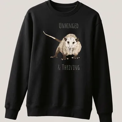 Sketch illustration of an opossum with the text 'UNHINGED & THRIVING' in earthy colors — printed on a unisex sweatshirt