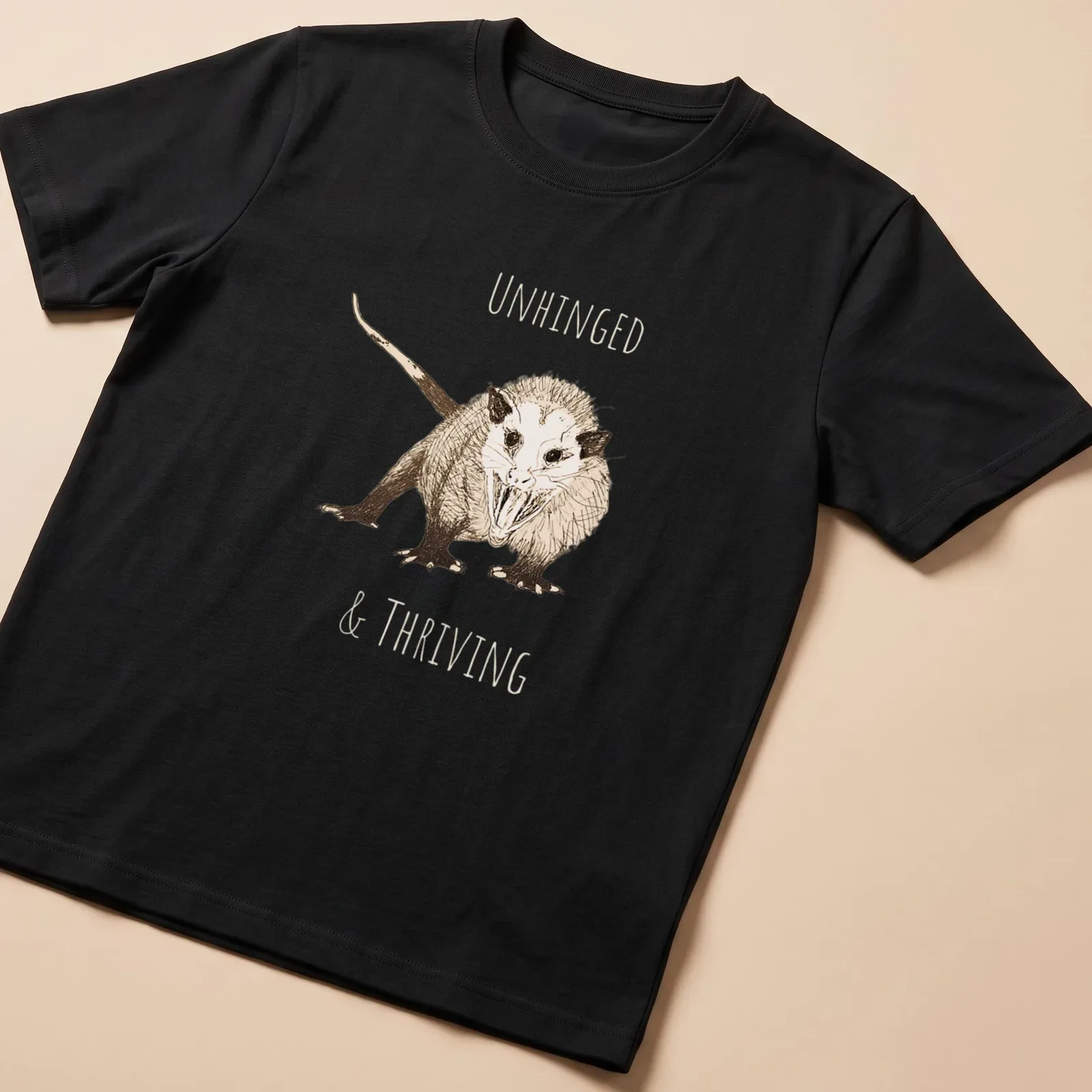 Sketch illustration of an opossum with the text 'UNHINGED & THRIVING' in earthy colors — printed on a unisex t-shirt