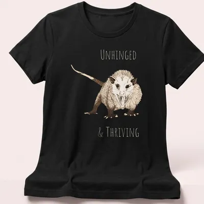 Sketch illustration of an opossum with the text 'UNHINGED & THRIVING' in earthy colors — printed on a women's shirt