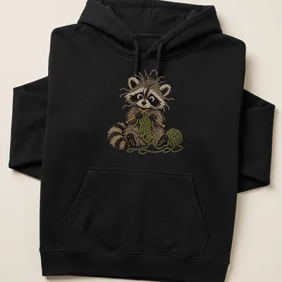 Whimsical raccoon knitting in faux cross stitch style, featuring brown and green colors — printed on a unisex hoodie