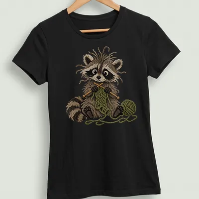 Whimsical raccoon knitting in faux cross stitch style, featuring brown and green colors — printed on a women's shirt