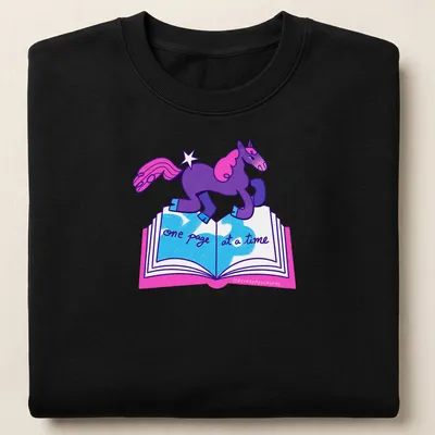 Colorful cartoon unicorn with text 'one page at a time' in purple, pink, and blue — printed on a unisex sweatshirt